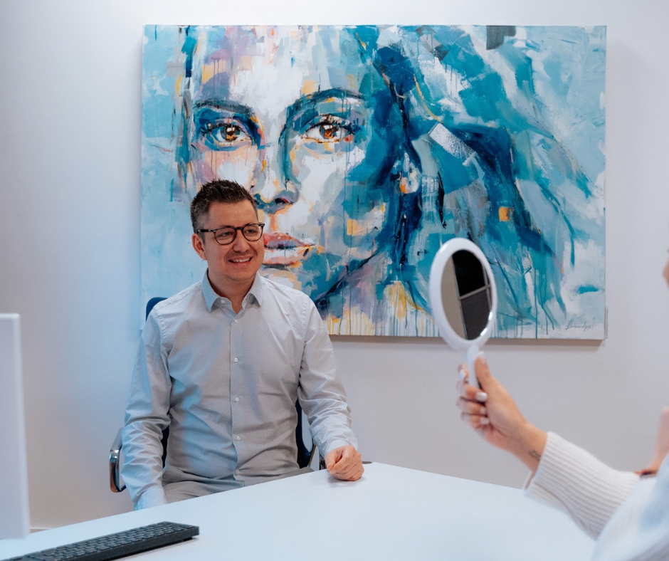 Arzt sitzt lachend am Schreibtisch gegenüber sitzt Frau mit Handspiegel